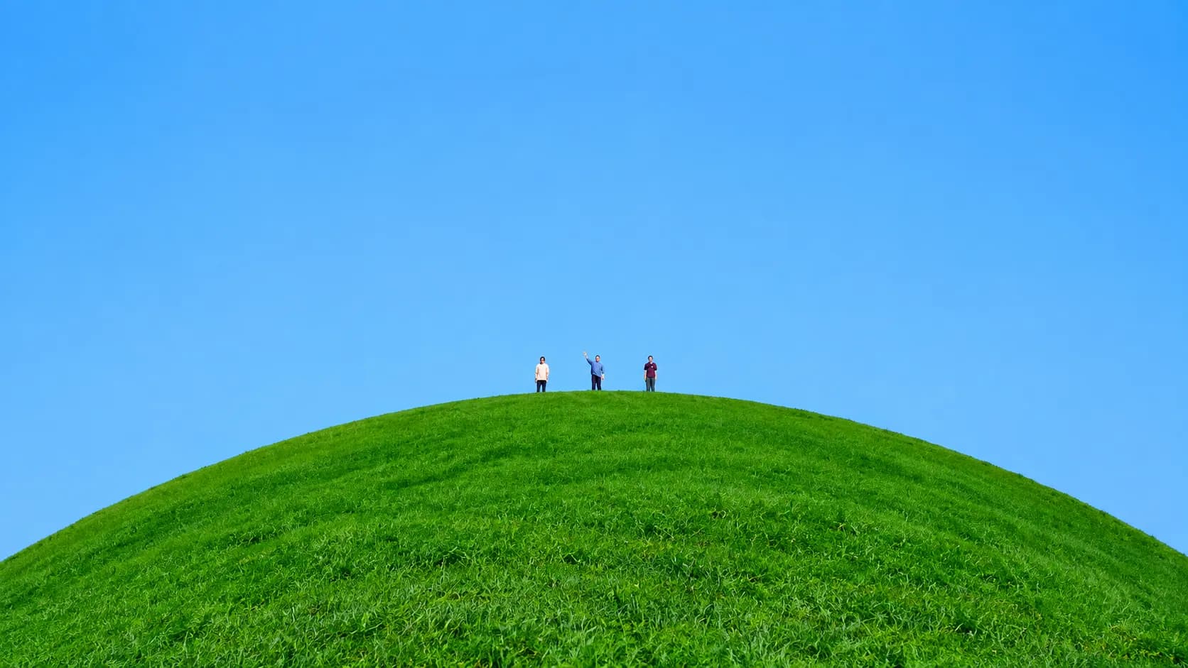 Drei Menschen stehen auf einem grünen Hügel unter blauem Himmel